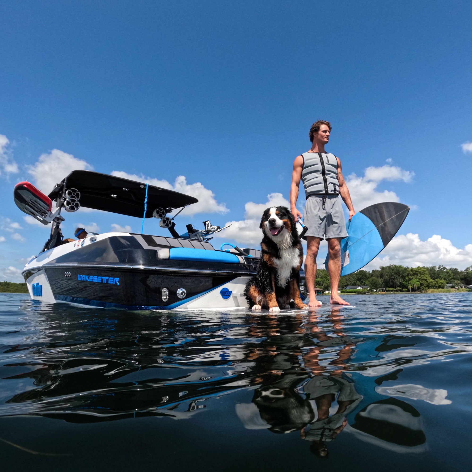 A man and his dog standing on a SWELL Wakesurf boat in the water.